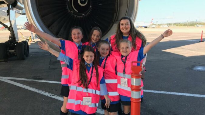 Five school girls and one adult wearing pink safety vests standing in front of a commercial airplane's jet engine on the tarmac